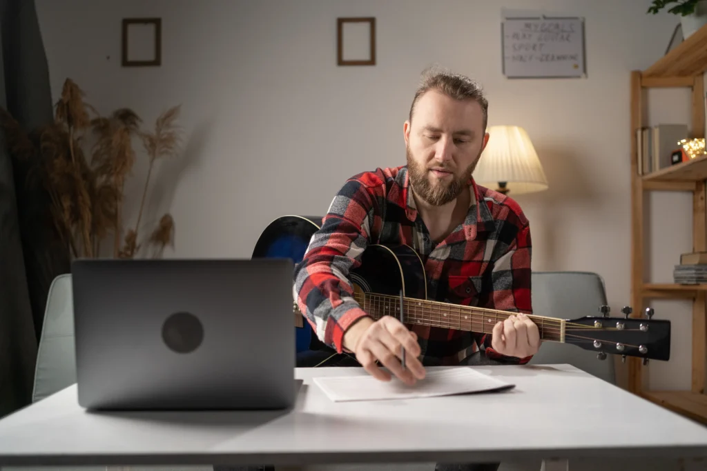 Young composer man composing song using guitar and laptop at home writing notes.